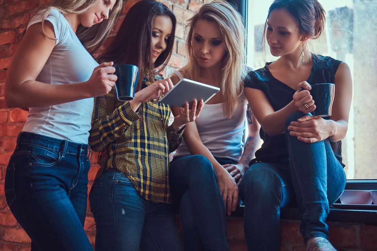 Group of female friends in casual clothes discussing while looking something on a digital tablet in a room with loft interior.